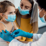 Little girl with protective face mask getting a vaccine while being with her mother at pediatrician's.