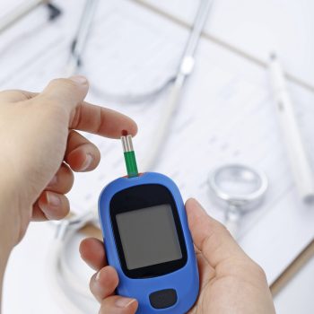 Hand holding a blood glucose meter measuring blood sugar, the background is a stethoscope and chart file